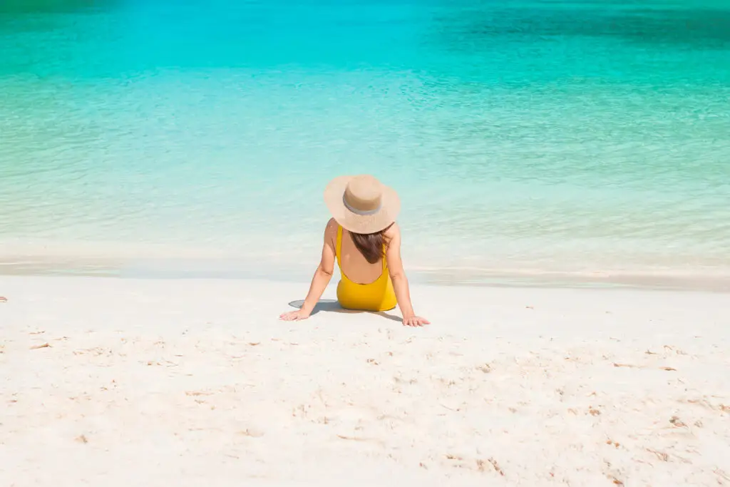 Person wearing a straw hat and yellow swimsuit sitting on white sand facing turquoise water