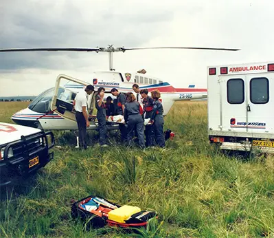 Emergency medical team transferring a patient from a helicopter to an ambulance in a grassy field, with Europ Assistance branding visible on the ambulance and helicopter