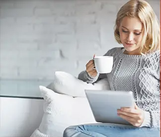Person sitting on a white couch holding a white coffee mug in one hand and a tablet in the other, wearing a light gray knitted sweater