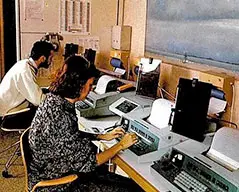 Two people working at desks equipped with vintage computer terminals and teleprinters in an office setting, with papers and technical equipment arranged on the workstations