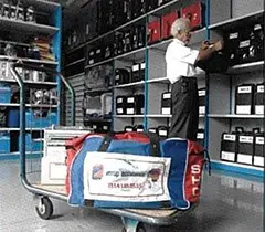 Person organizing black bags on shelves in a storage room, with a trolley in the foreground carrying a large red and blue Europ Assistance duffel bag and other items