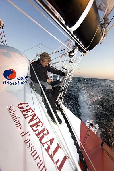 Person sitting on the deck of a racing sailboat with Generali and Europ Assistance branding, navigating through open sea under clear skies