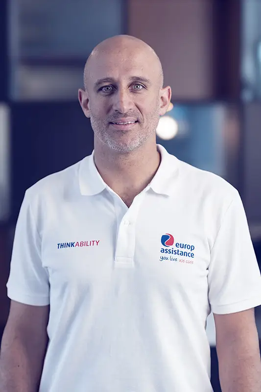 A middle-aged man with a shaved head and close-cropped facial stubble looks directly at the camera with a neutral, friendly expression. He wears a white polo shirt featuring two logos—"ThinkAbility" (with the "Ability" highlighted in red) on the left chest and the "Europ Assistance – you live we care" logo on the right. The background is softly blurred, keeping attention on his face and attire.