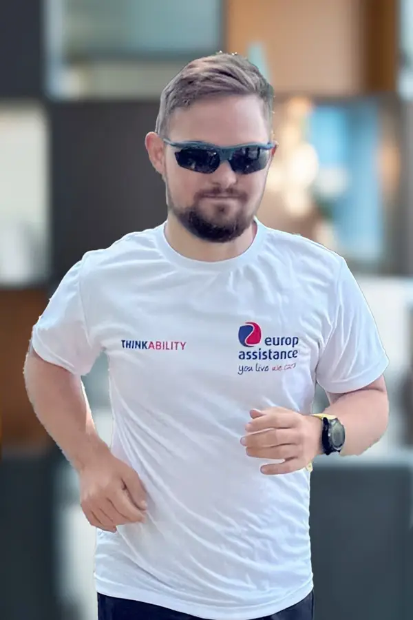 A casually dressed man is walking or jogging forward. He’s wearing dark sunglasses and has short hair and a trimmed beard. His T-shirt is white with two visible logos: one on the left side of the chest says "ThinkAbility" (with the “Ability” highlighted in red), and the other—on the right—reads "Europ Assistance — you live we care." He also sports a wristwatch or fitness tracker on his left wrist, and the background appears softly blurred, suggesting perhaps a shallow depth of field, which keeps focus on him.