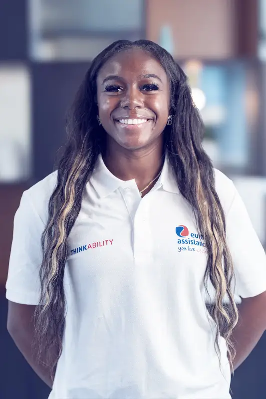 A young Black woman stands smiling warmly at the camera. She wears a white polo shirt with logos—“ThinkAbility” on the left and “Europ Assistance — you live we care.” on the right. She has long, wavy hair and subtle makeup, including long eyelashes and hoop earrings; she also sports a small nose ring. The background is softly out of focus (likely indoors), keeping the focus on her bright, friendly expression.