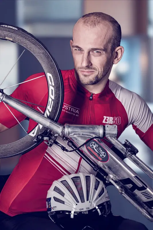 Person wearing a red and white cycling jersey holding a road bike over the shoulder, with a white helmet positioned in front