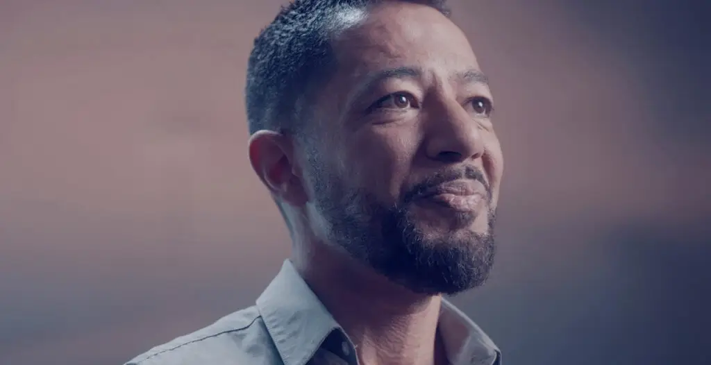 Person wearing a light gray collared shirt, shown from the shoulders up against a softly lit background