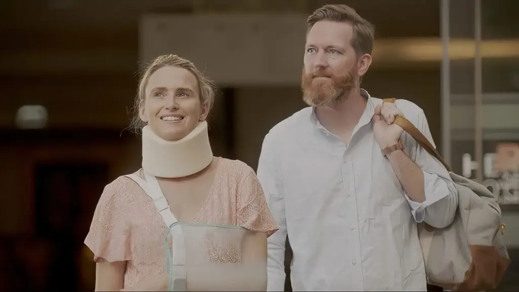 Two people standing indoors, one wearing a medical neck brace and arm sling, the other carrying a shoulder bag