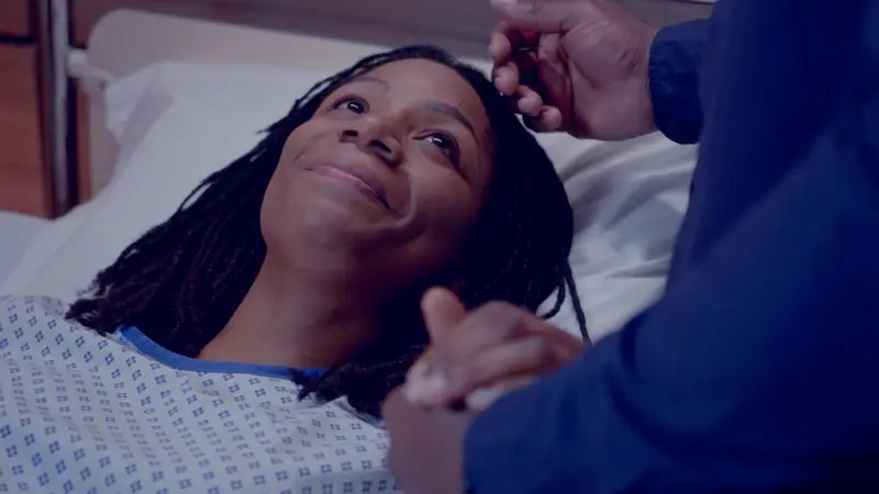 Person lying in a hospital bed wearing a patterned medical gown, with another person holding their hand